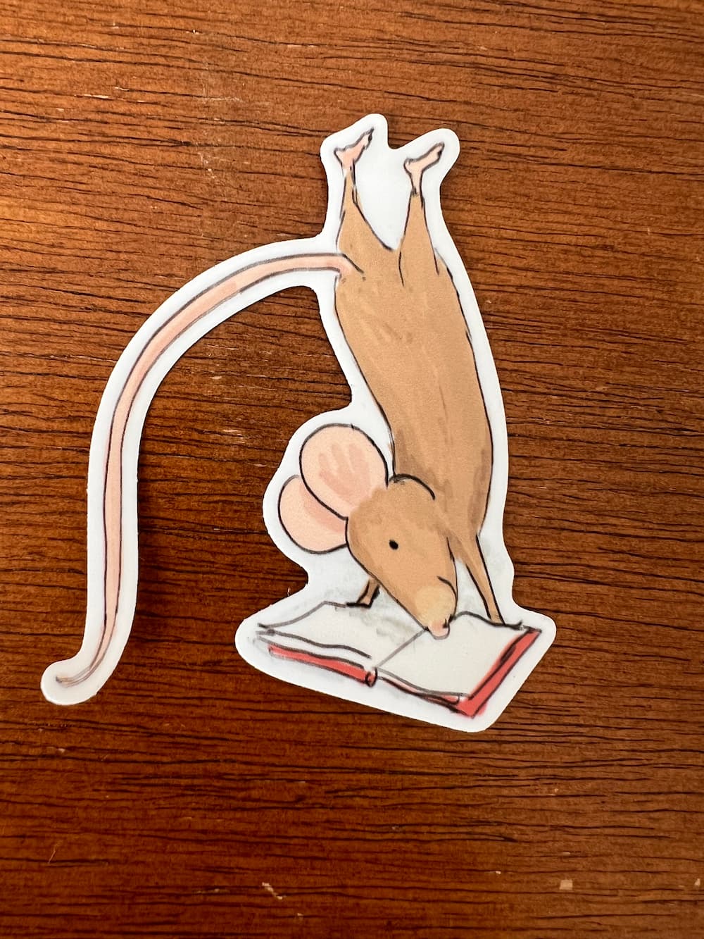 A die-cut sticker featuring an illustration of a brown mouse hanging upside down over an open book, placed on a brown wooden surface.