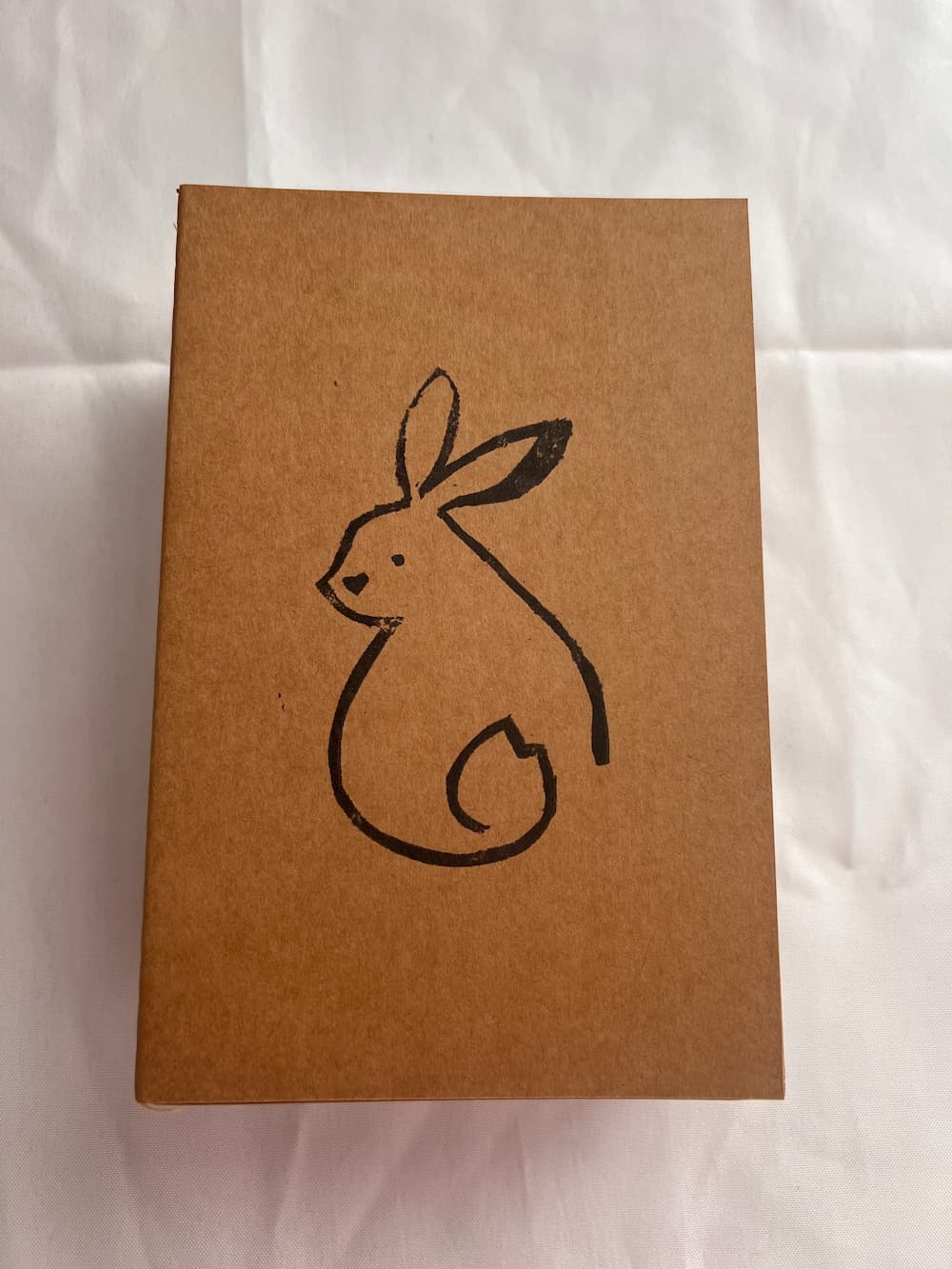 A square brown kraft paper box with a simple black line drawing of a rabbit on the front, photographed against a white fabric background.