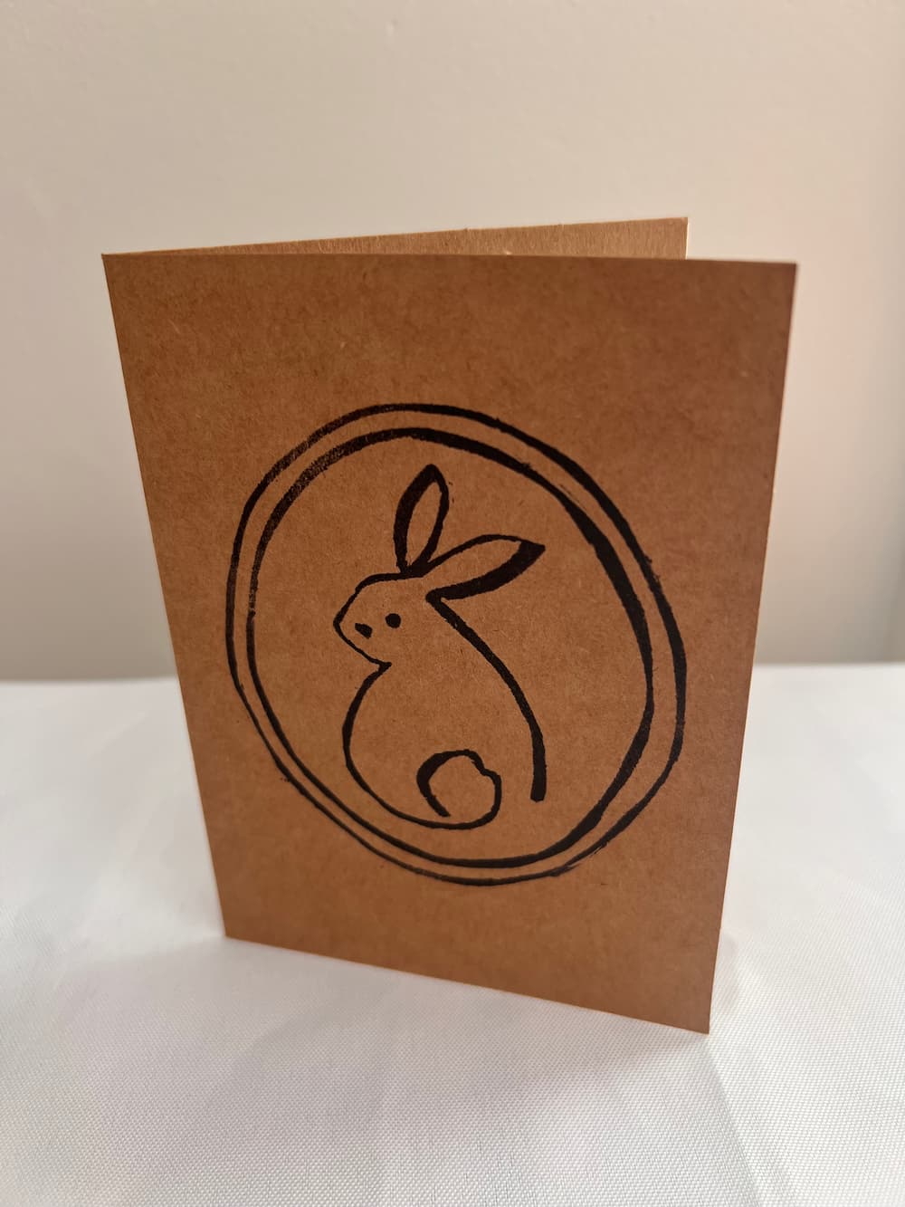 A square brown kraft paper card with a black hand-drawn rabbit inside a circle, standing upright on a white surface against a light beige background.
