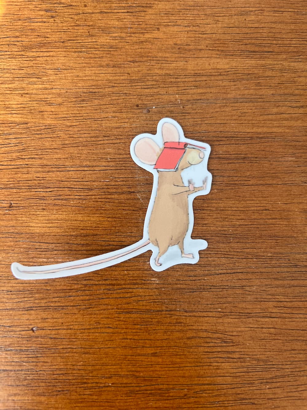 A die-cut sticker of a cartoon mouse wearing a red visor, placed on a brown wooden surface.