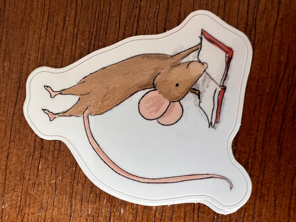 A die-cut sticker featuring an illustration of a brown mouse climbing a red ladder, placed on a brown wooden surface.